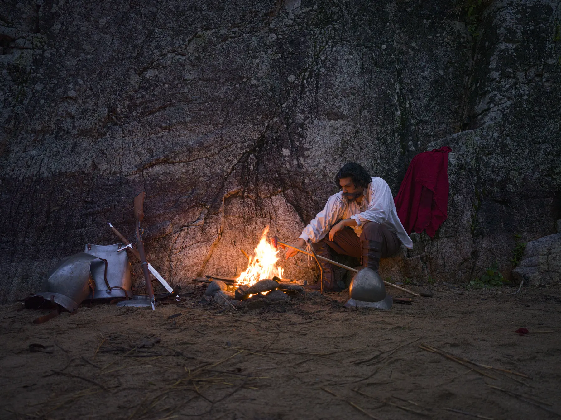 A figure tends a fire against a rock face at dusk